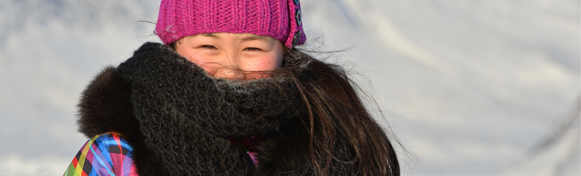 Photo d'une jeune personne portant un bonnet en tricot rose et un foulard marron foncé qui couvre la majeure partie de son visage. Elle se tient devant un arrière-plan enneigé.